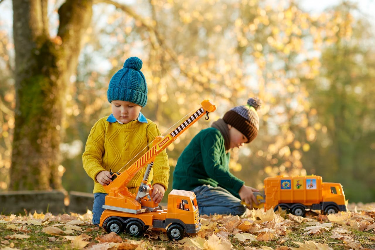 Two kids playing with toy trucks in a scenic park during autumn, surrounded by colorful leaves.