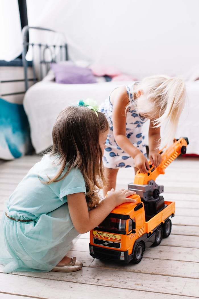 Menu Two young girls playing with a toy truck on the floor of a bright and cozy bedroom.