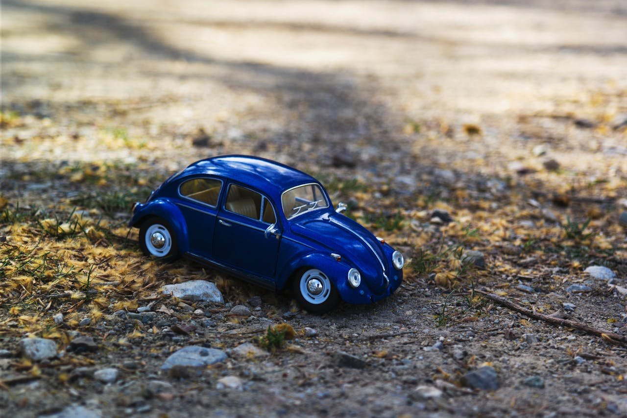 A miniature blue Volkswagen Beetle toy car on a sunlit gravel path, surrounded by nature.
